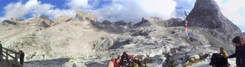 Panoramica dal terrazzo del rifugio. Nel tavolo al centro &egrave; seduto il gruppo del coro Sosat, che ride e scherza preparandosi per la serata e il giorno successivo giocando a carte e godendosi il bel paesaggio. Sulla destra, la Iv coccola un bel cagnone arrivato fin quass&ugrave; e molto socievole con tutti